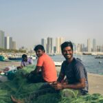 Fishermen repairing nets by the seashore with Sharjah city skyline in the background.