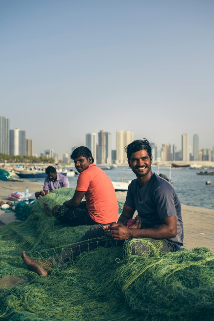Fishermen repairing nets by the seashore with Sharjah city skyline in the background.